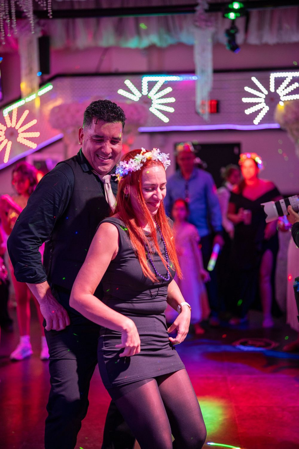 Couple dancing together at a vibrant nightclub party with colorful lights and guests in the background.