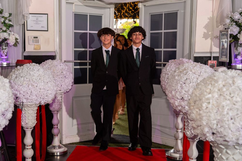 Two young men in black suits walk a red carpet flanked by white floral arrangements at a formal prom event.