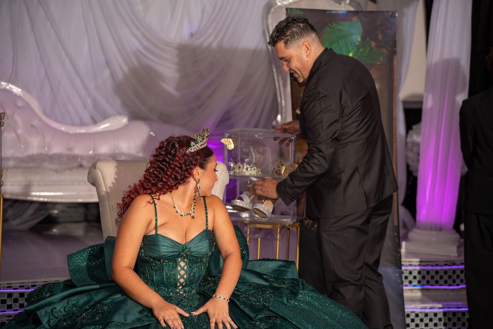 Young woman in emerald green quinceañera gown seated as man in black suit presents gift at formal party.