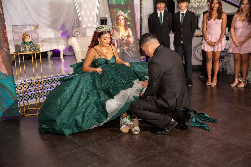 A young woman in a green quinceañera gown receives the last doll ceremony while guests watch at her elegant celebration.