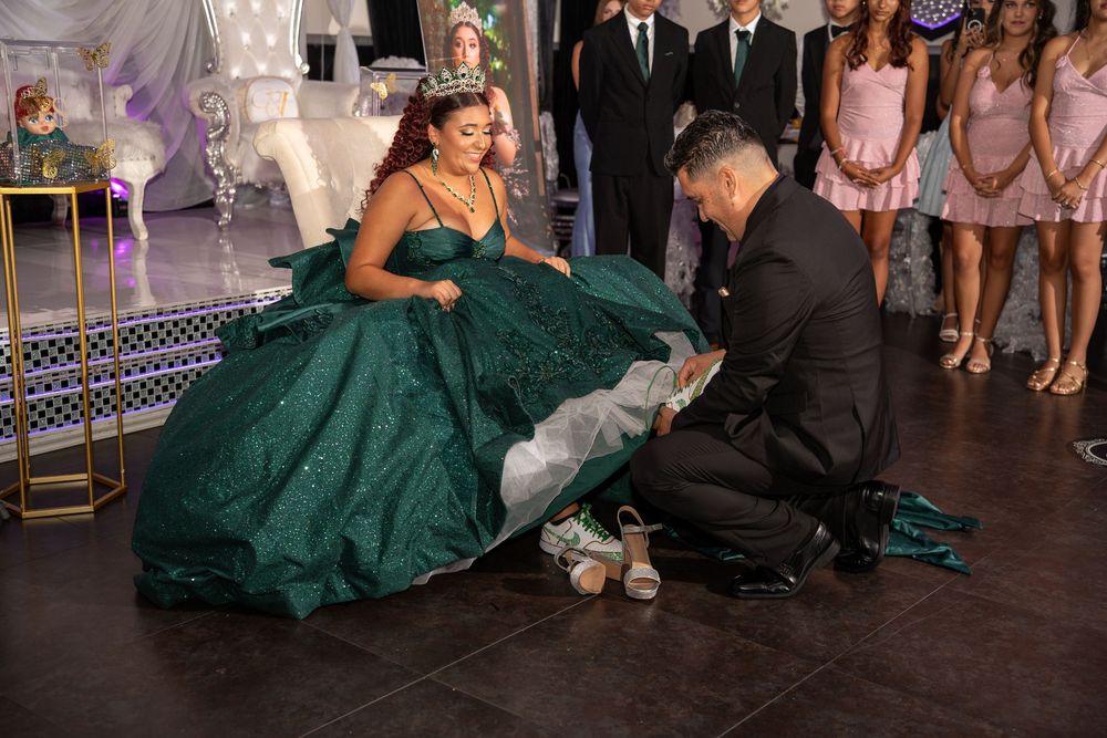 A young woman in a green quinceañera gown wearing a crown while a man kneels to place her shoes during the celebration.