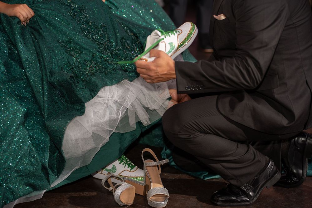 A man in a black suit helps a bride in a green sequin gown put on green Converse sneakers at a wedding reception.