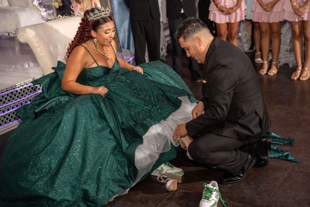 Man kneeling to place shoes on a girl in a sparkling green quinceañera ball gown during her celebration.
