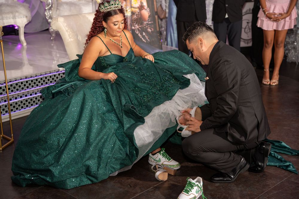 A young woman in a green quinceañera gown receives sneakers from a man kneeling before her at a formal celebration.