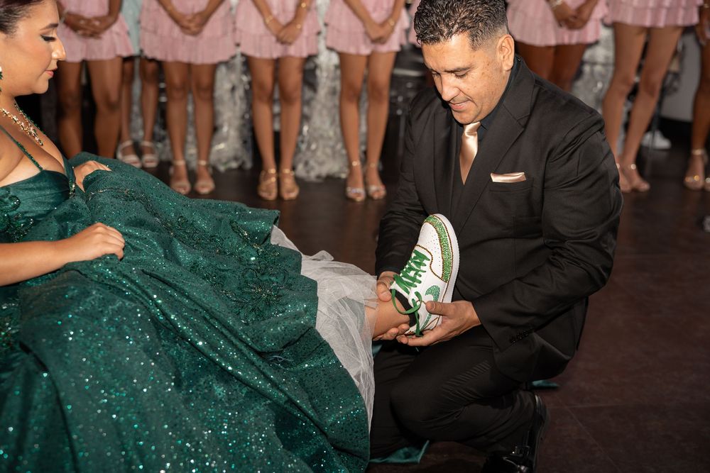 Man in black suit places green sneaker on woman in emerald sequin gown during quinceañera last dance ceremony.