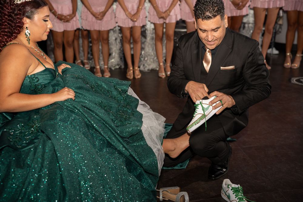 Man in black suit puts green sneakers on woman in sparkling emerald ball gown at formal event.