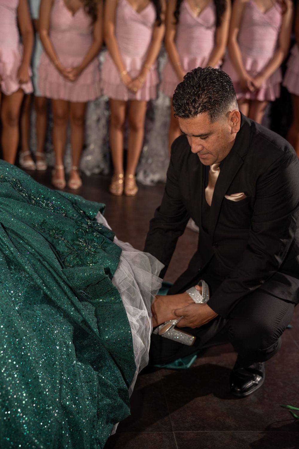 Man in black suit kneeling beside a green sequin gown at a quinceañera, with bridesmaids in pink dresses behind.