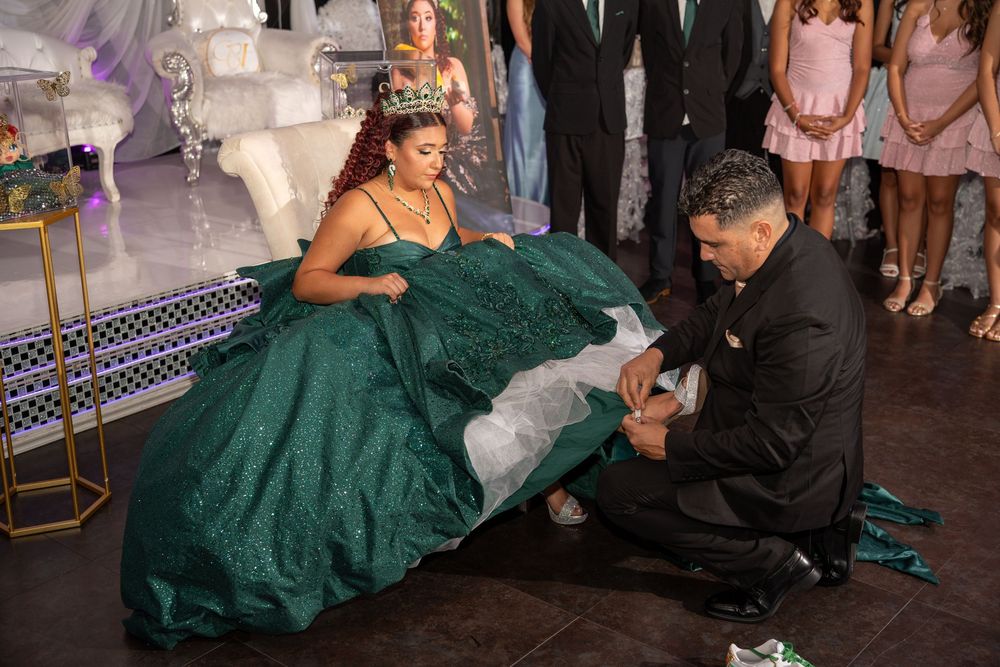 A man kneels to place a shoe on a young woman in a green quinceañera ball gown at an elegant celebration.