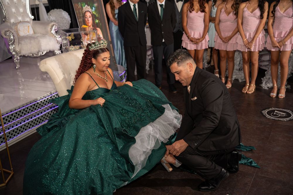 A man kneels to place a shoe on a quinceañera girl in a green ballgown at her celebration.