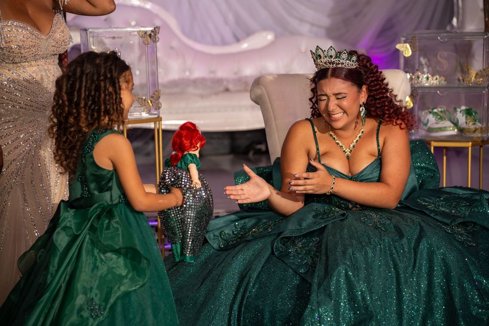 Young girl presents a red rose to a smiling quinceañera in a green ball gown wearing a crown at her celebration.