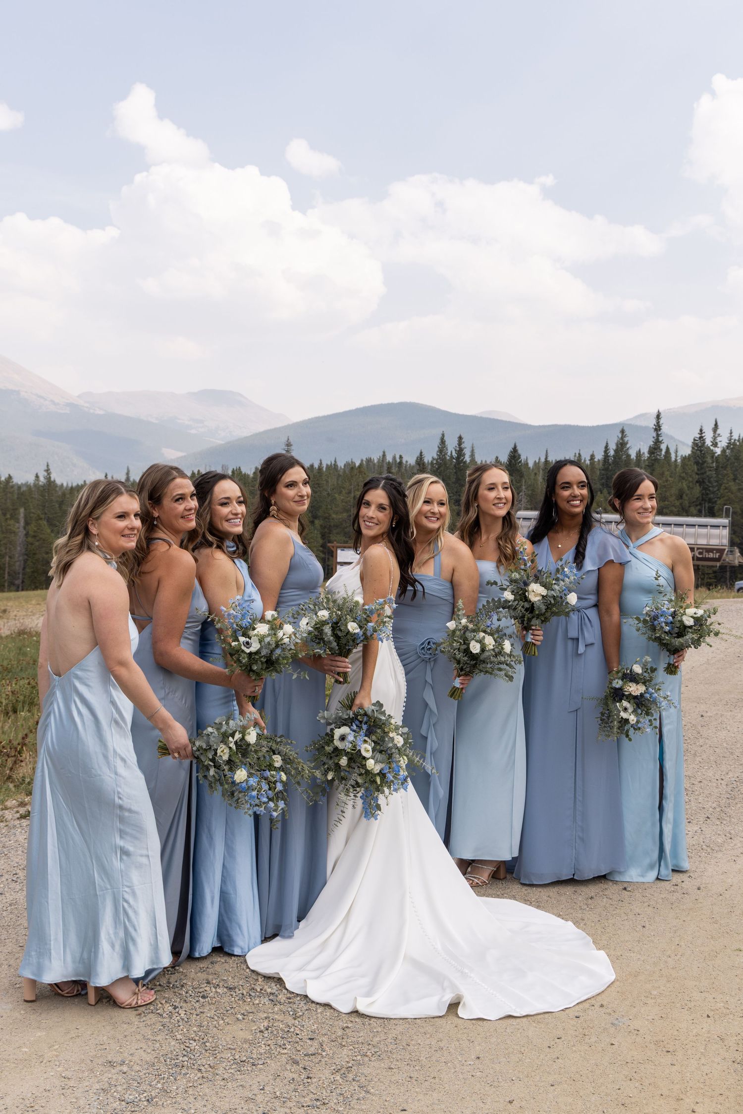Bride in white gown with bridesmaids in dusty blue dresses holding white bouquets, posing outdoors with mountain backdrop.
