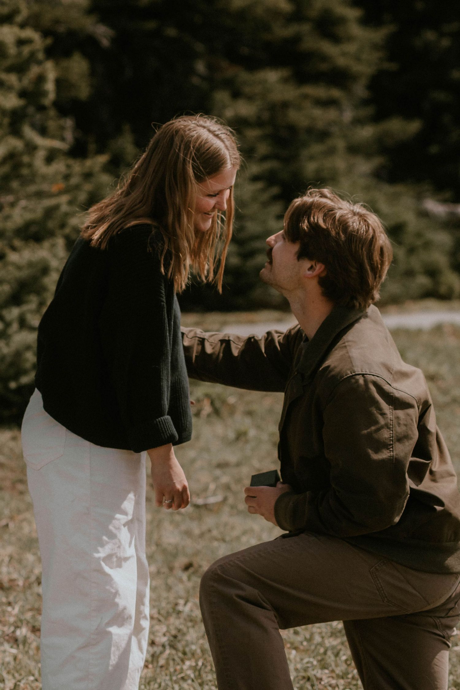 Man kneeling on one knee proposing to a smiling woman outdoors with evergreen trees in the background.