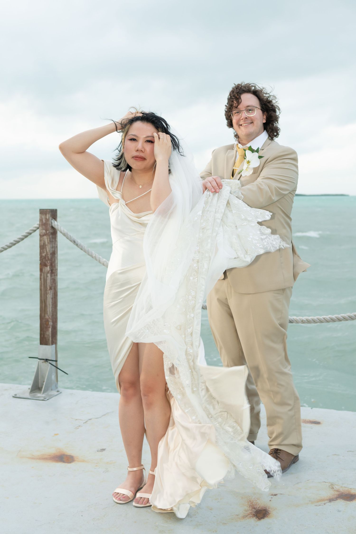 Bride and groom pose by the ocean on their wedding day, wind blowing her white gown and veil.