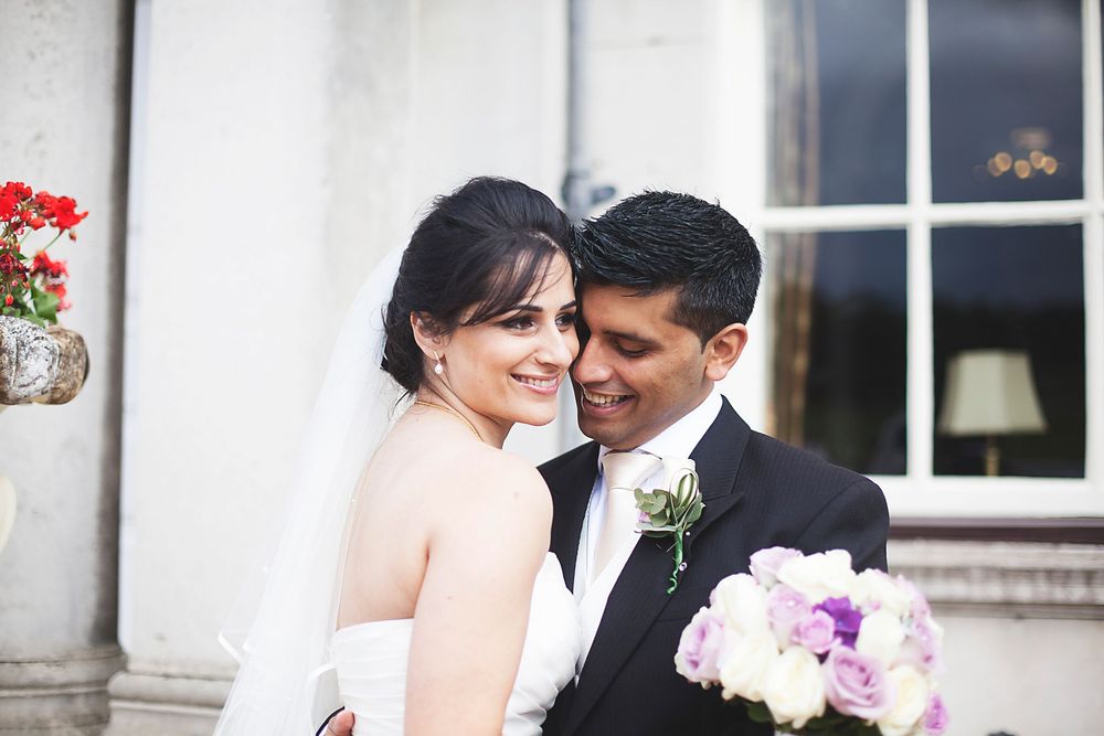 A smiling bride in a white veil holds a purple bouquet beside her groom in a black tuxedo with a floral boutonniere.