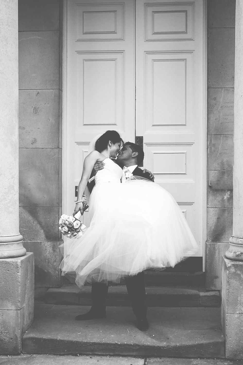 Black and white photo of a groom lifting his bride in a wedding dress, sharing a kiss in front of elegant double doors.