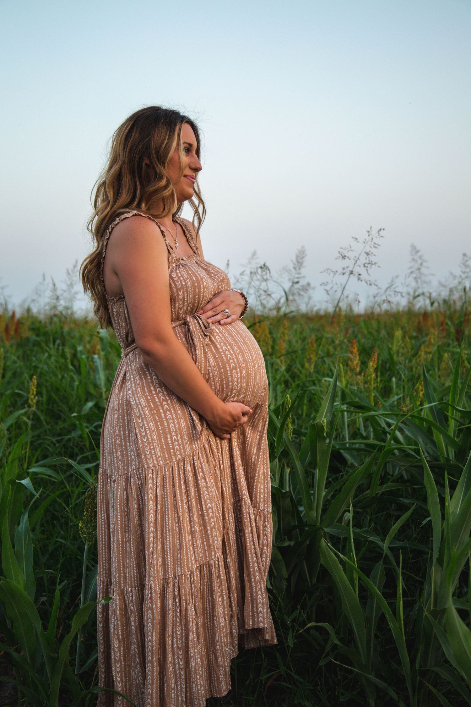 A pregnant woman in a pink maxi dress cradles her baby bump in a lush green cornfield at dusk.