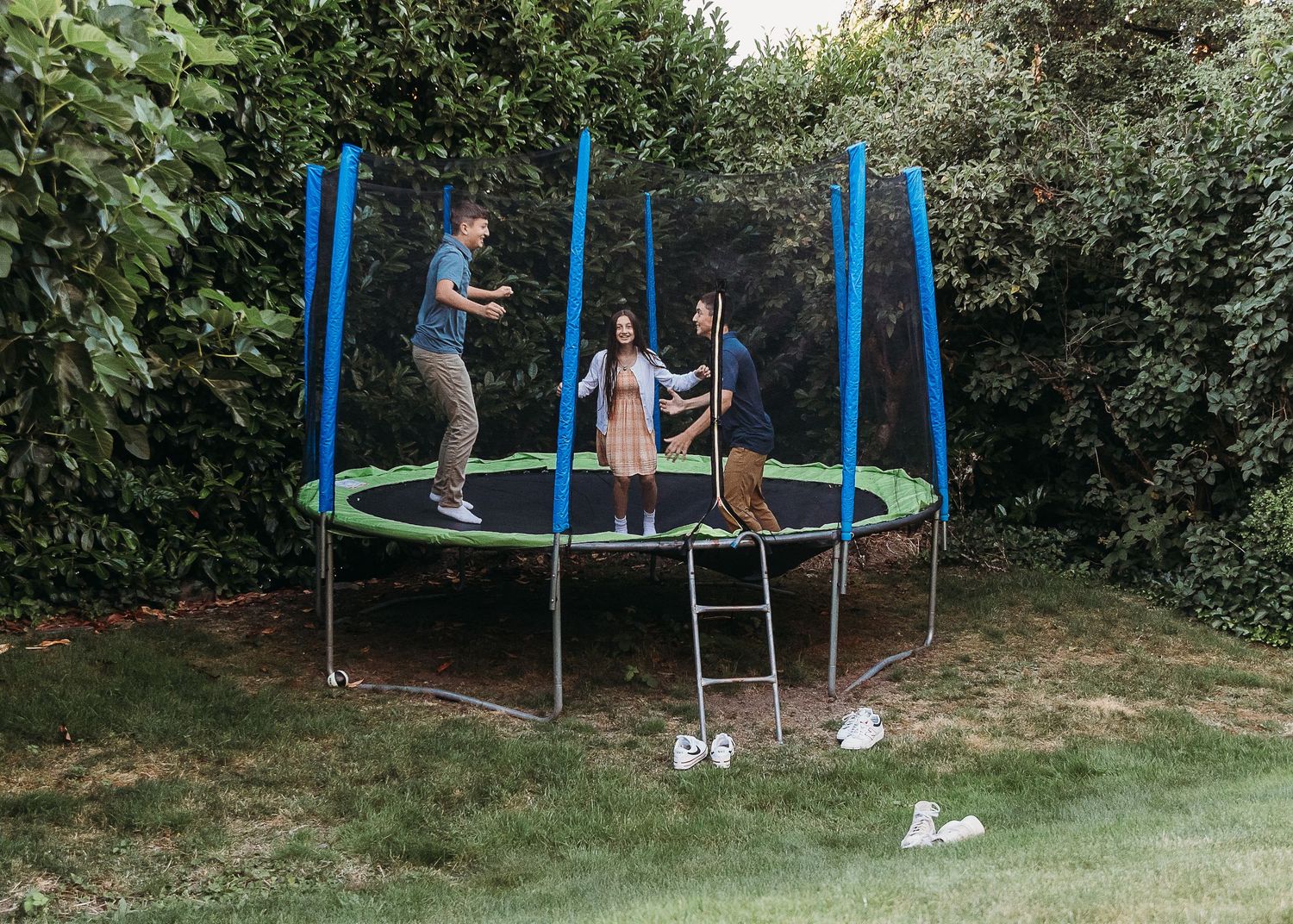 Three people enjoying a large outdoor trampoline with safety net enclosure in a lush green backyard.