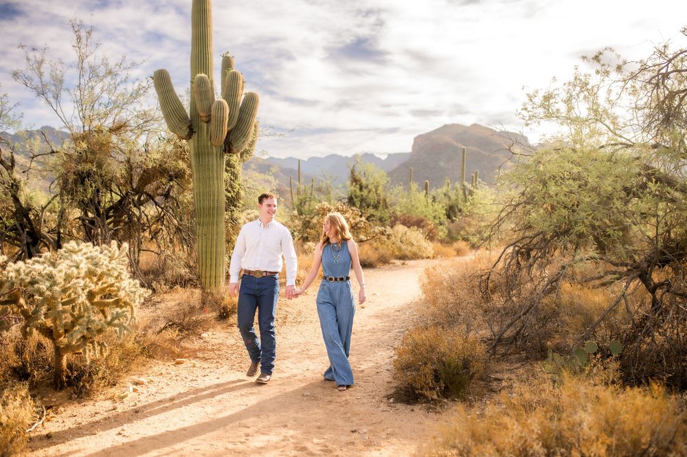 Happy couple holding hands walking on a desert trail surrounded by saguaro cacti and mountains.