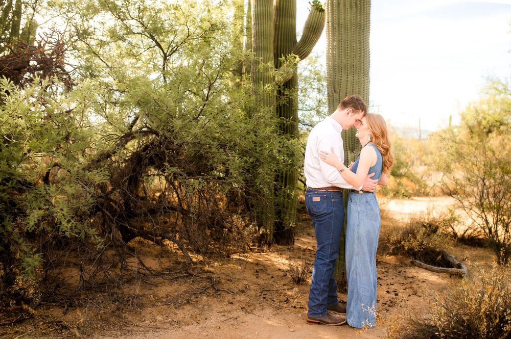 Couple sharing a romantic kiss on a desert path surrounded by saguaro cacti and lush greenery.