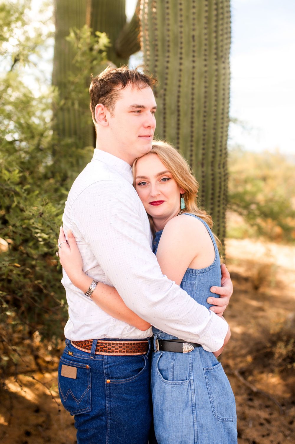 Young couple embracing warmly beside a tall saguaro cactus in the Arizona desert landscape.