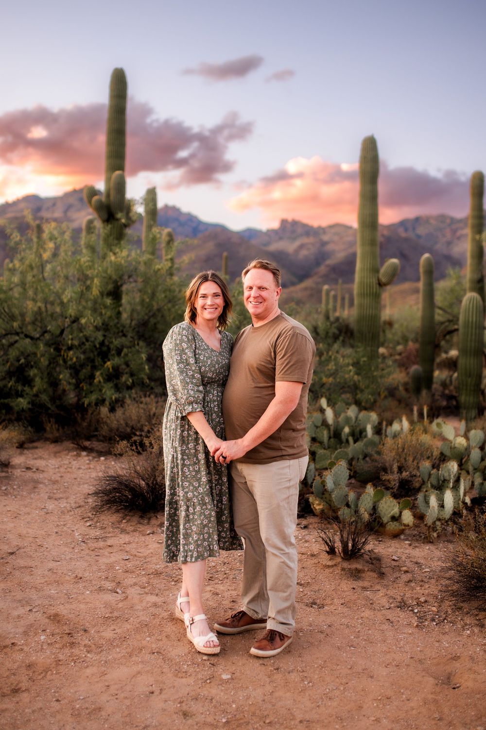Smiling couple holding hands in a desert setting with saguaro cacti and a colorful sunset sky.