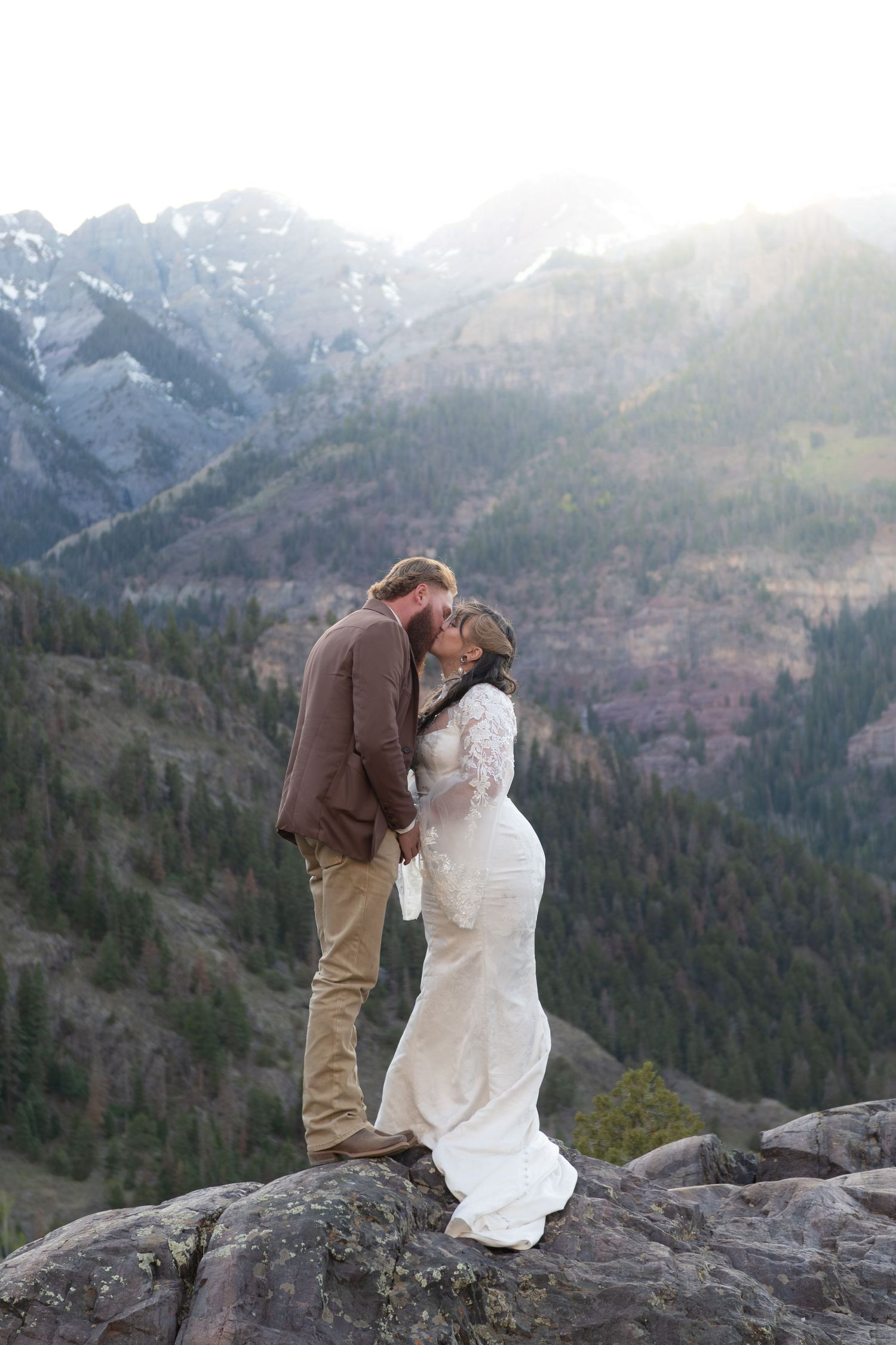 A bride and groom share a romantic kiss on a rocky mountain overlook with snow-capped peaks and pine forests behind them.