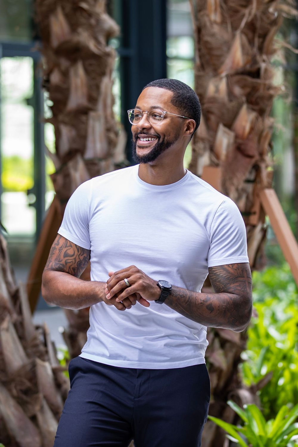 Man in white t-shirt smiles outdoors with palm trees in background.