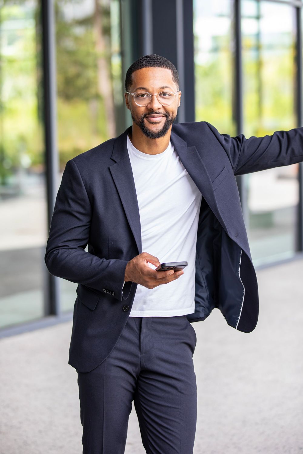 Professional in a navy suit and white t-shirt stands confidently with smartphone against window backdrop.