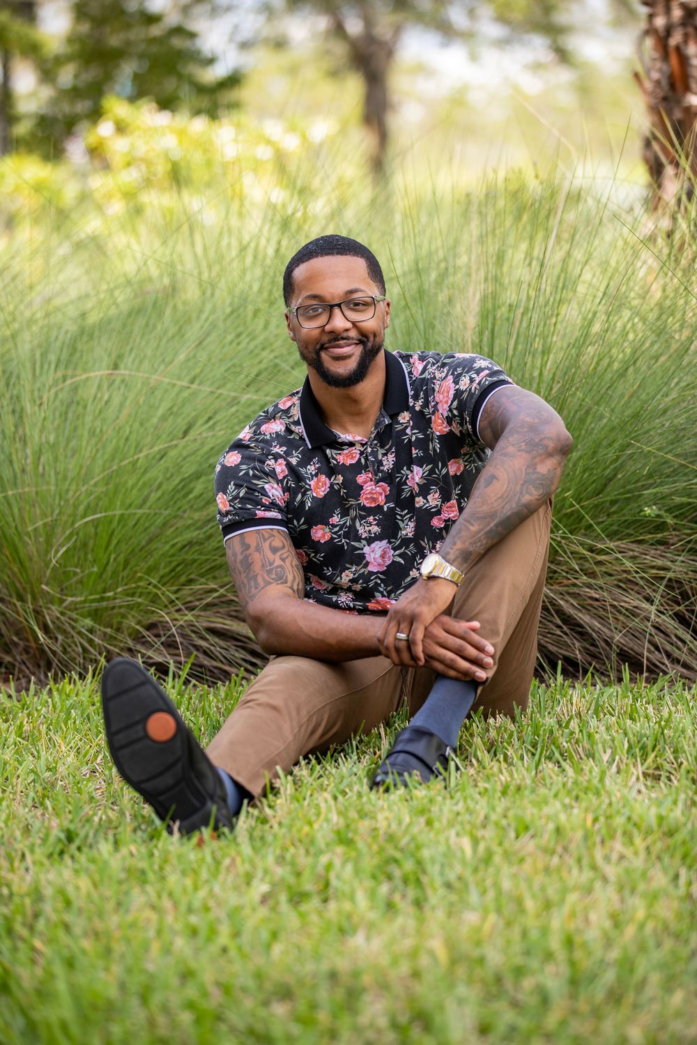 Person in floral shirt sitting on grass showing warm smile and casual style with glasses.