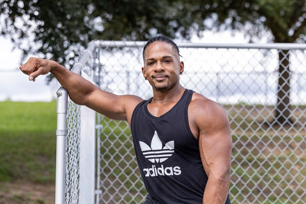 Muscular athlete in Adidas tank top poses confidently by chain link fence.