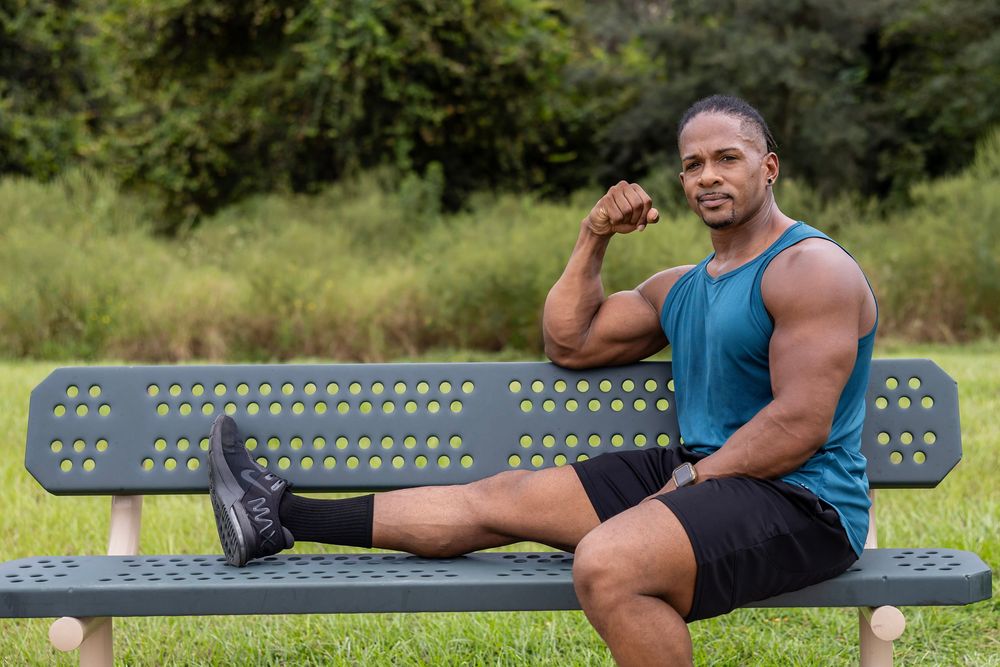 Muscular athlete in blue tank top relaxing on park bench during outdoor workout.