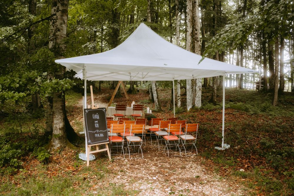White canopy tent with orange chairs set up in a forest clearing for an outdoor event.