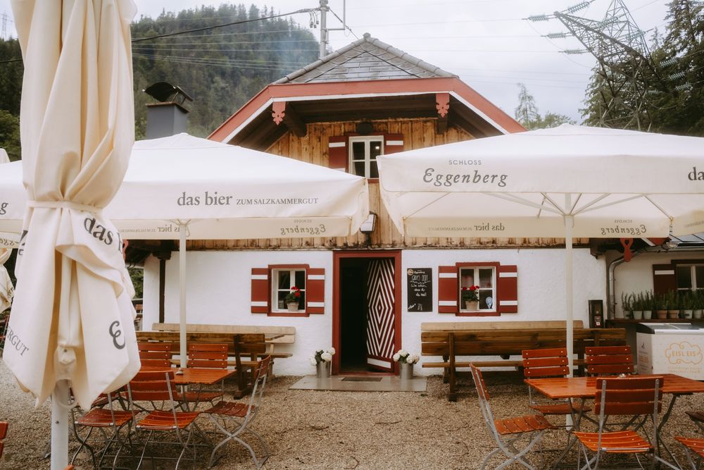 Eggenberg beer garden with white umbrellas, red and white alpine building, wooden chairs and tables on a rainy day.