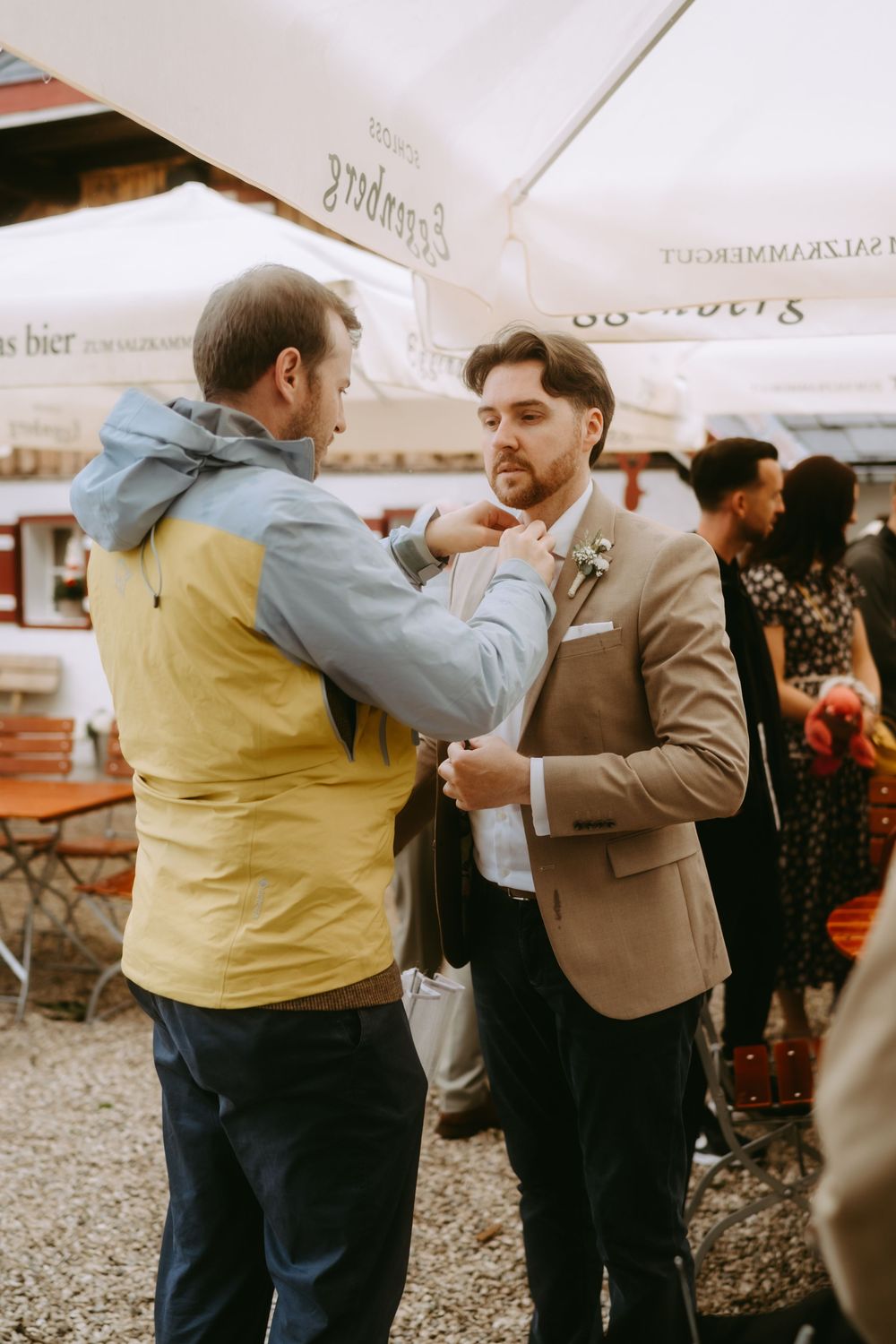 Groom in tan blazer gets bow tie adjusted by friend at outdoor wedding reception near beer garden.