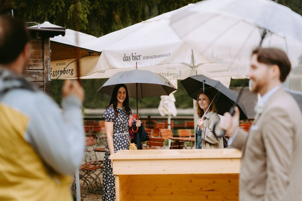 Two women holding umbrellas stand behind a wooden bar at an outdoor rainy event with white parasols.