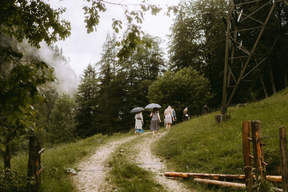 Group of people walking on a winding gravel path through lush green hills, some holding umbrellas on a misty day.