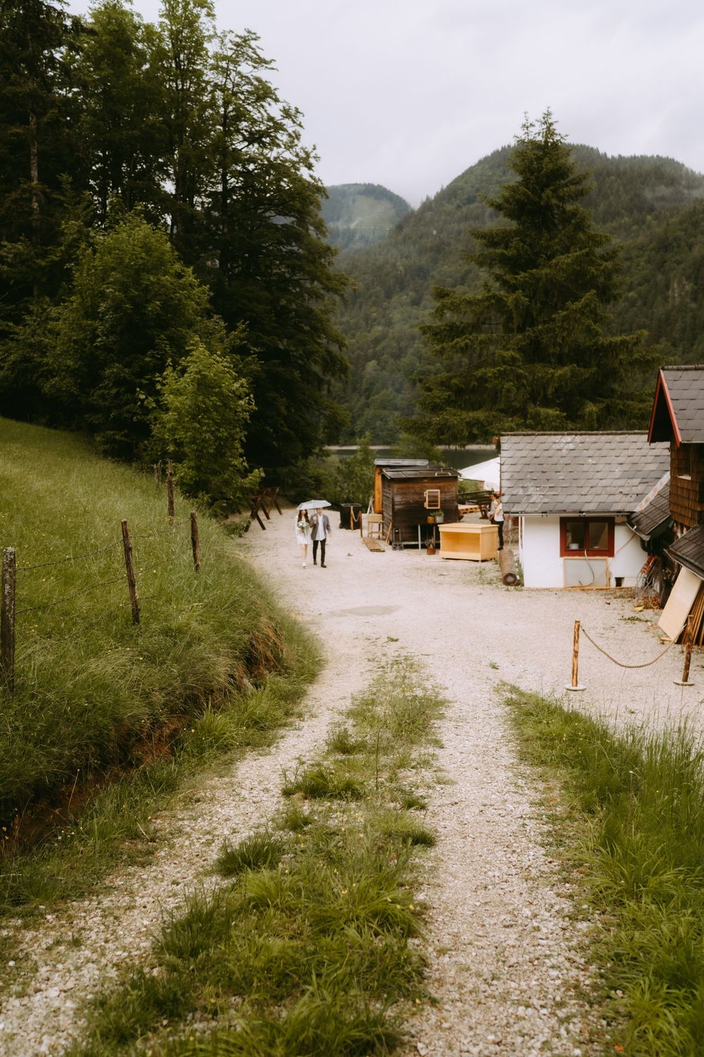 A rustic gravel path leads through an Alpine village with wooden cabins, lush green trees, and misty mountains in the background.