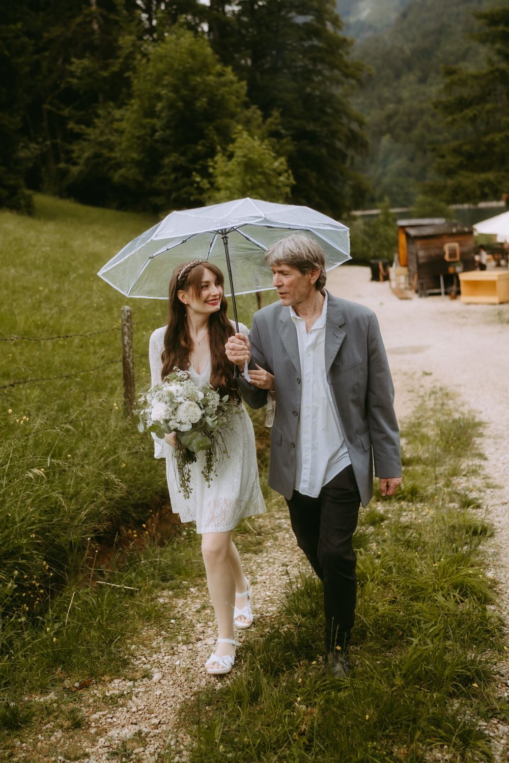 A smiling bride in a white dress holding a bouquet walks with a man under an umbrella on a rainy Alpine wedding day.