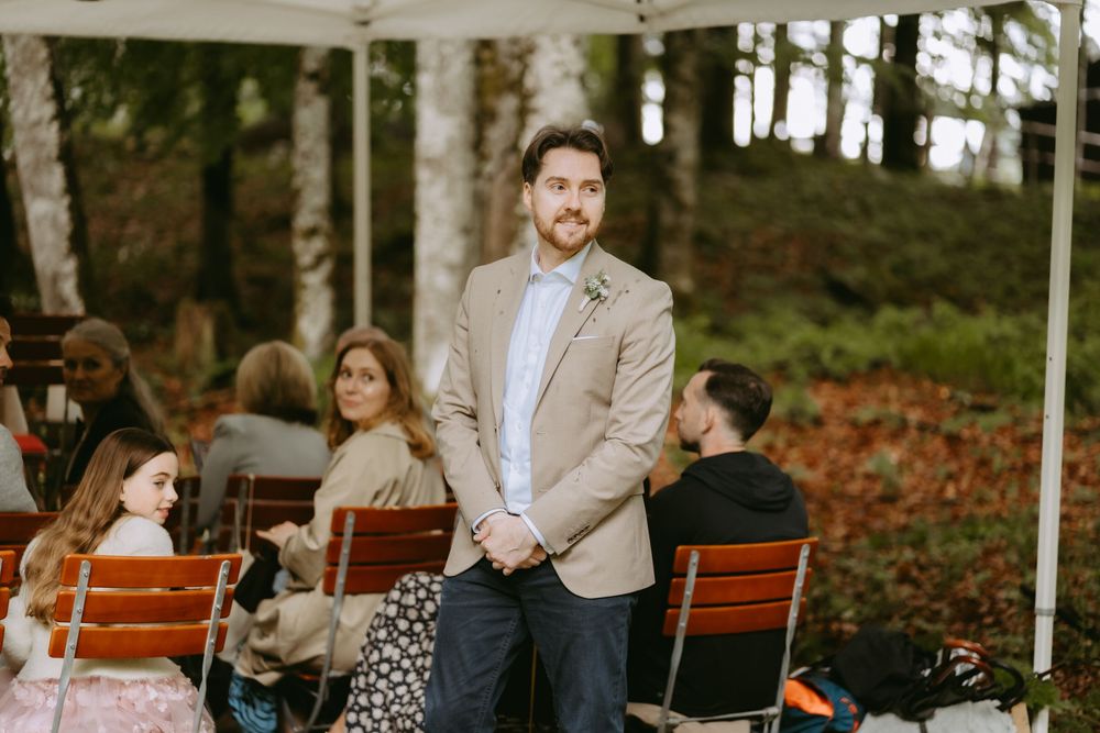 Groom in beige blazer stands outdoors at wedding ceremony with guests seated on benches among autumn trees.