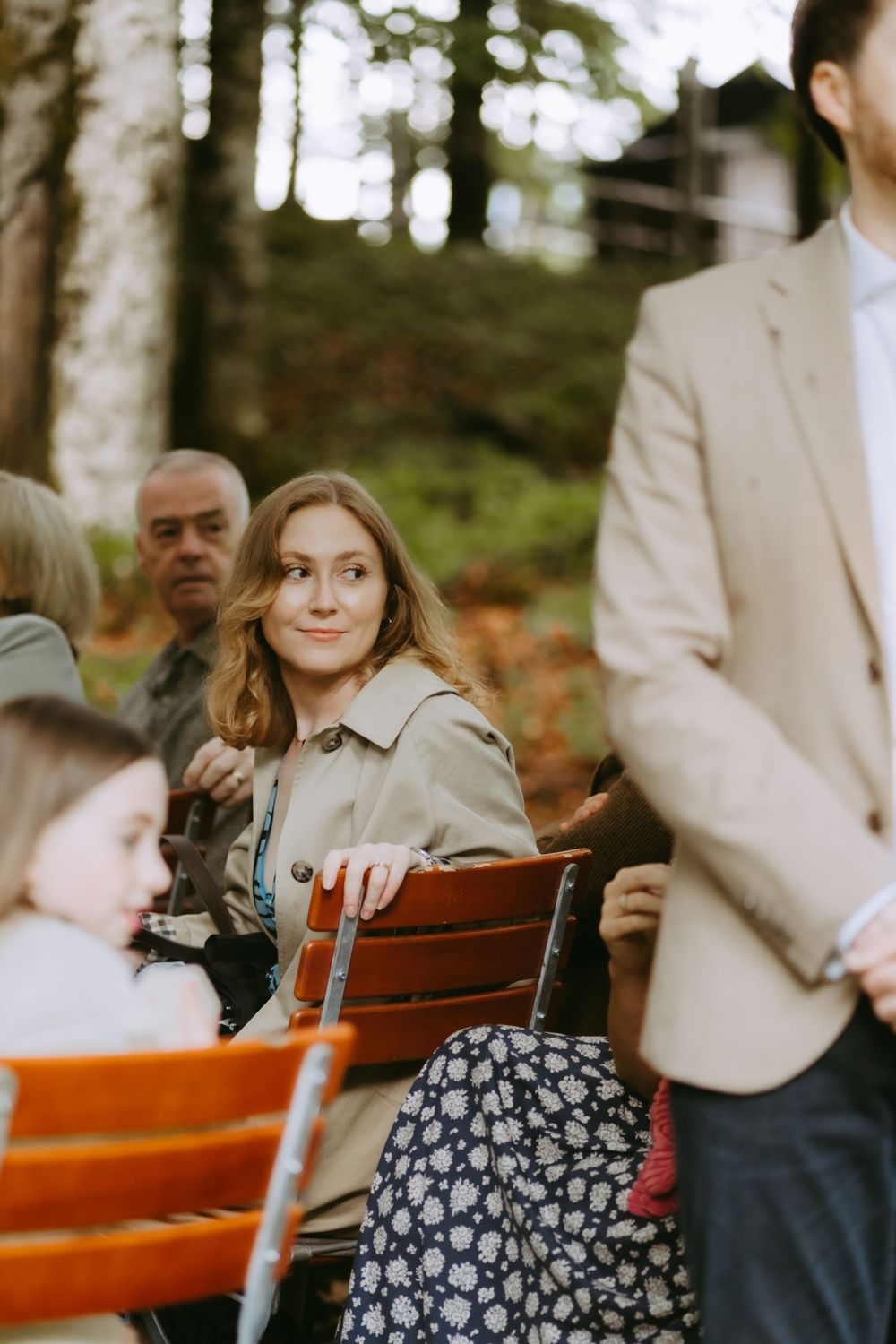 Wedding guest in trench coat sits on wooden bench outdoors among trees during ceremony.
