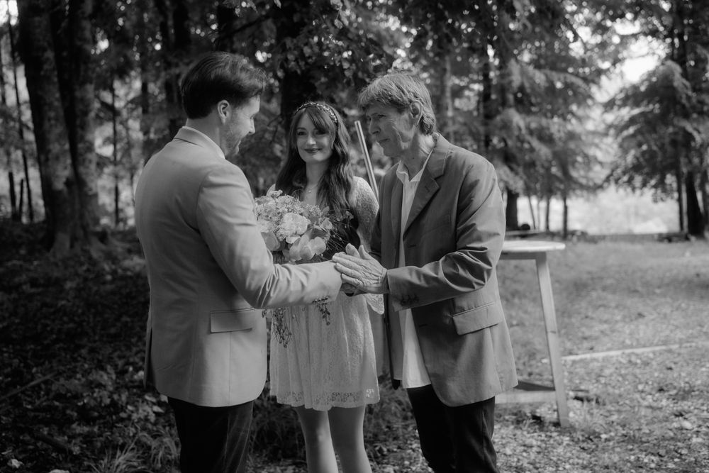 Black and white photo of a couple exchanging rings during an outdoor wedding ceremony in a forest setting.