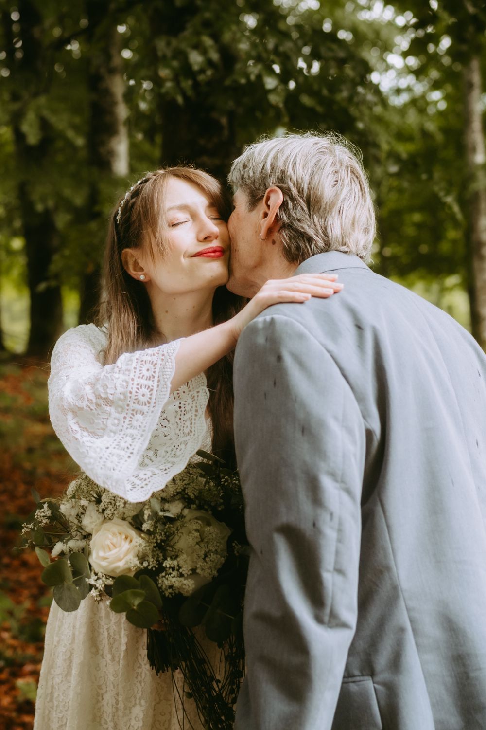 Bride in lace dress smiles as groom kisses her cheek in a sunlit forest wedding photo.
