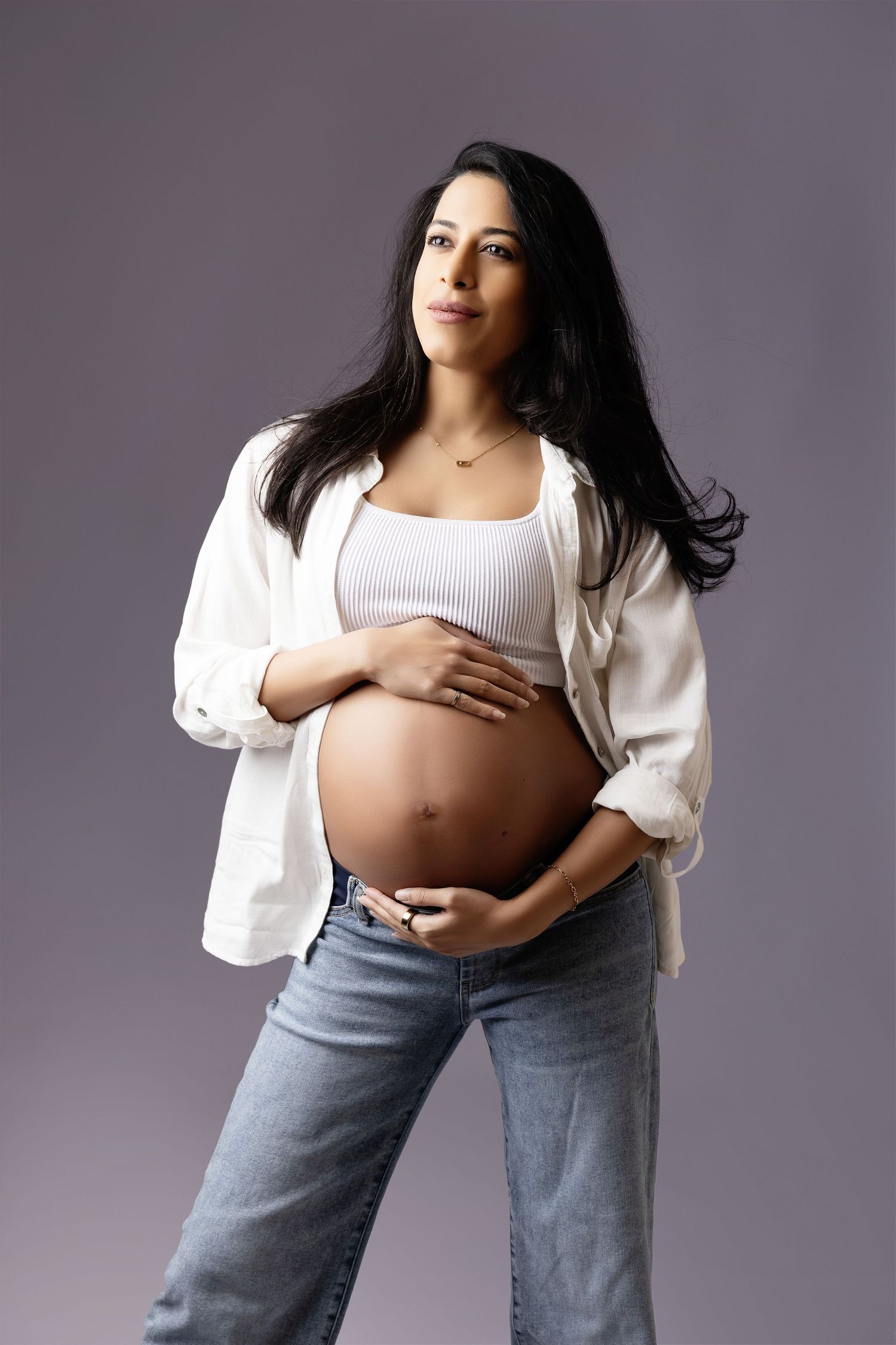 Pregnant woman in white shirt and jeans cradling baby bump, studio portrait on gray background.
