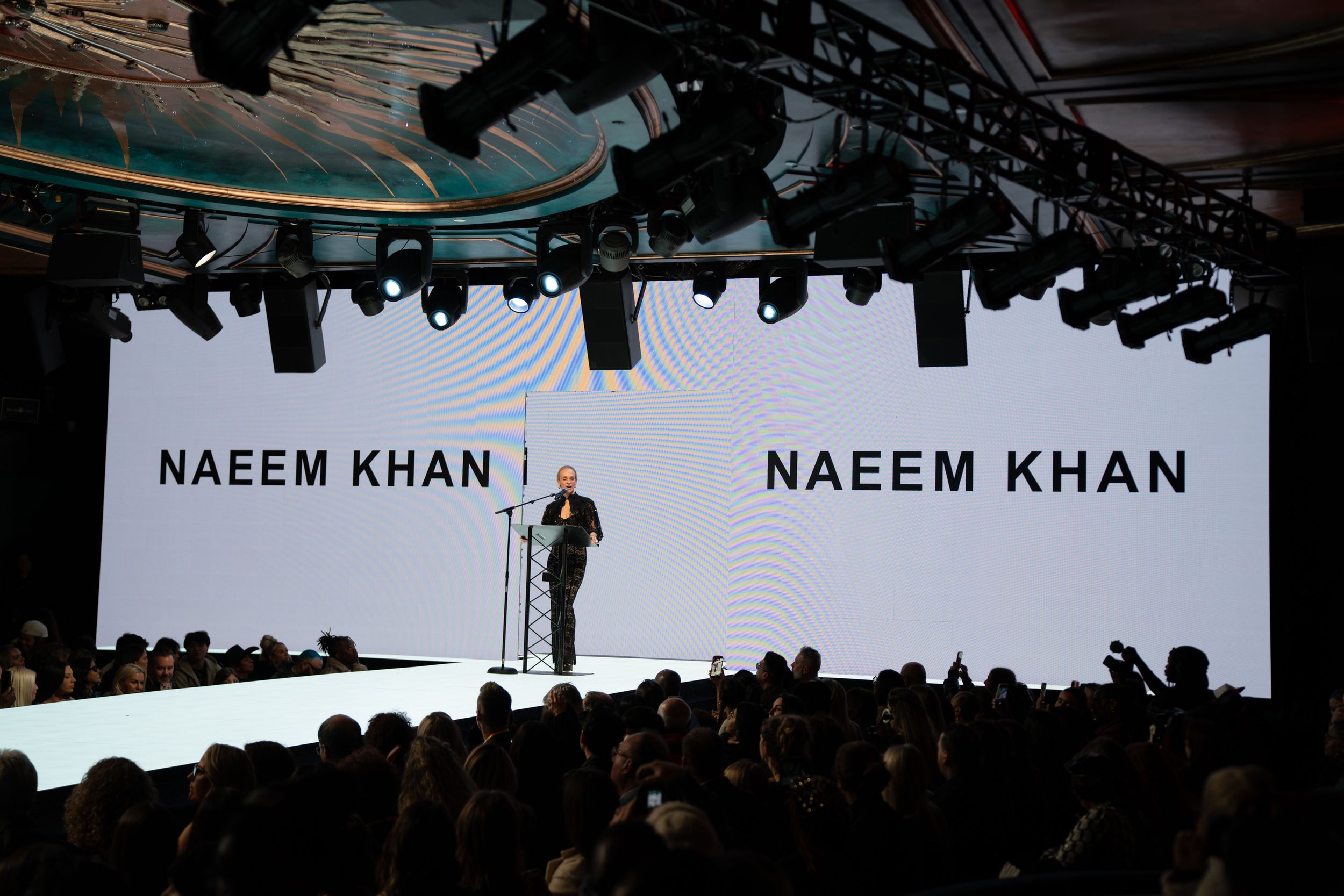 Naeem Khan fashion show with designer on stage before large LED screen displaying brand name at elegant runway event.