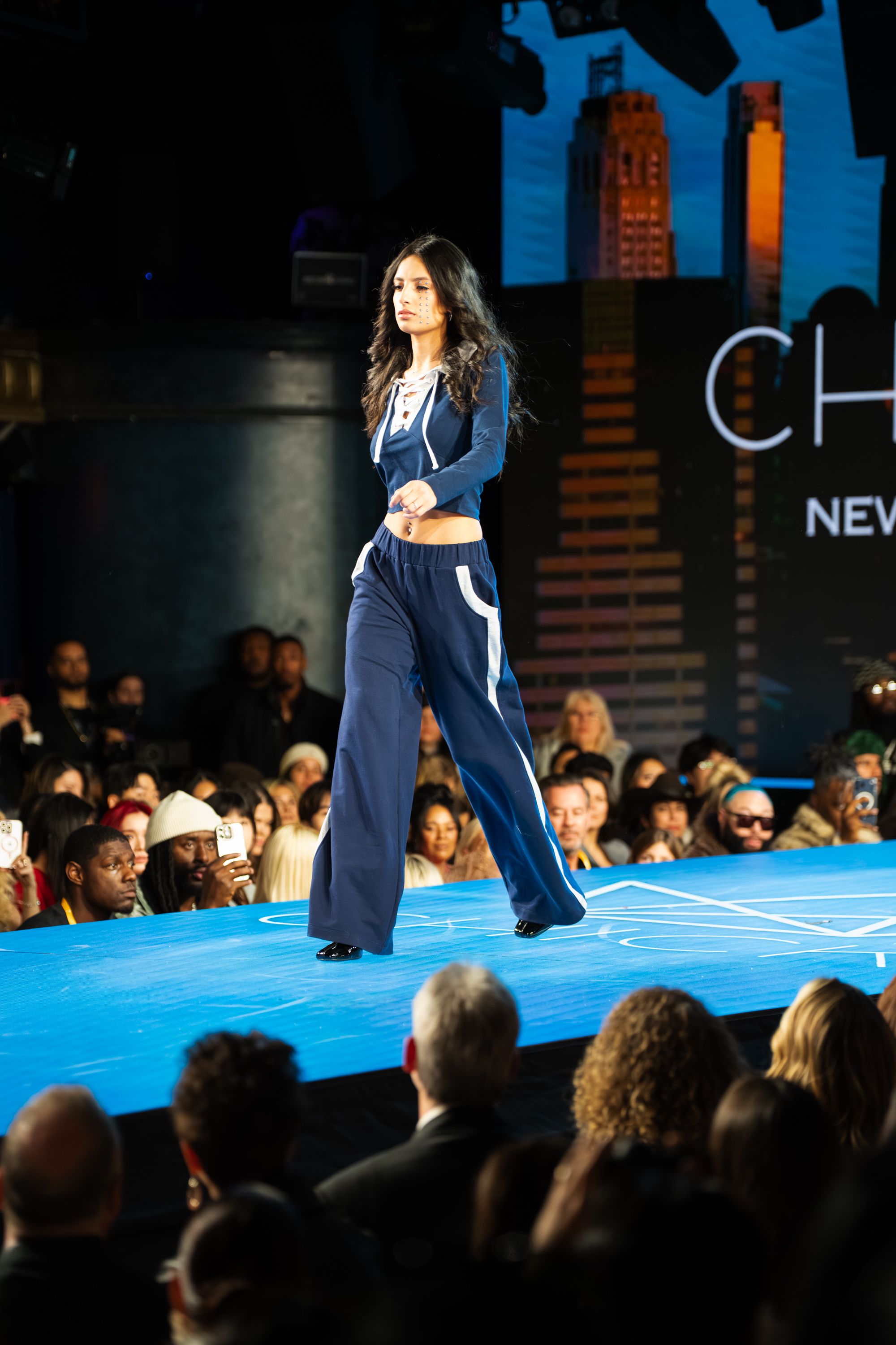 Model walks runway in navy blue wide-leg pants and crop jacket at a fashion show.