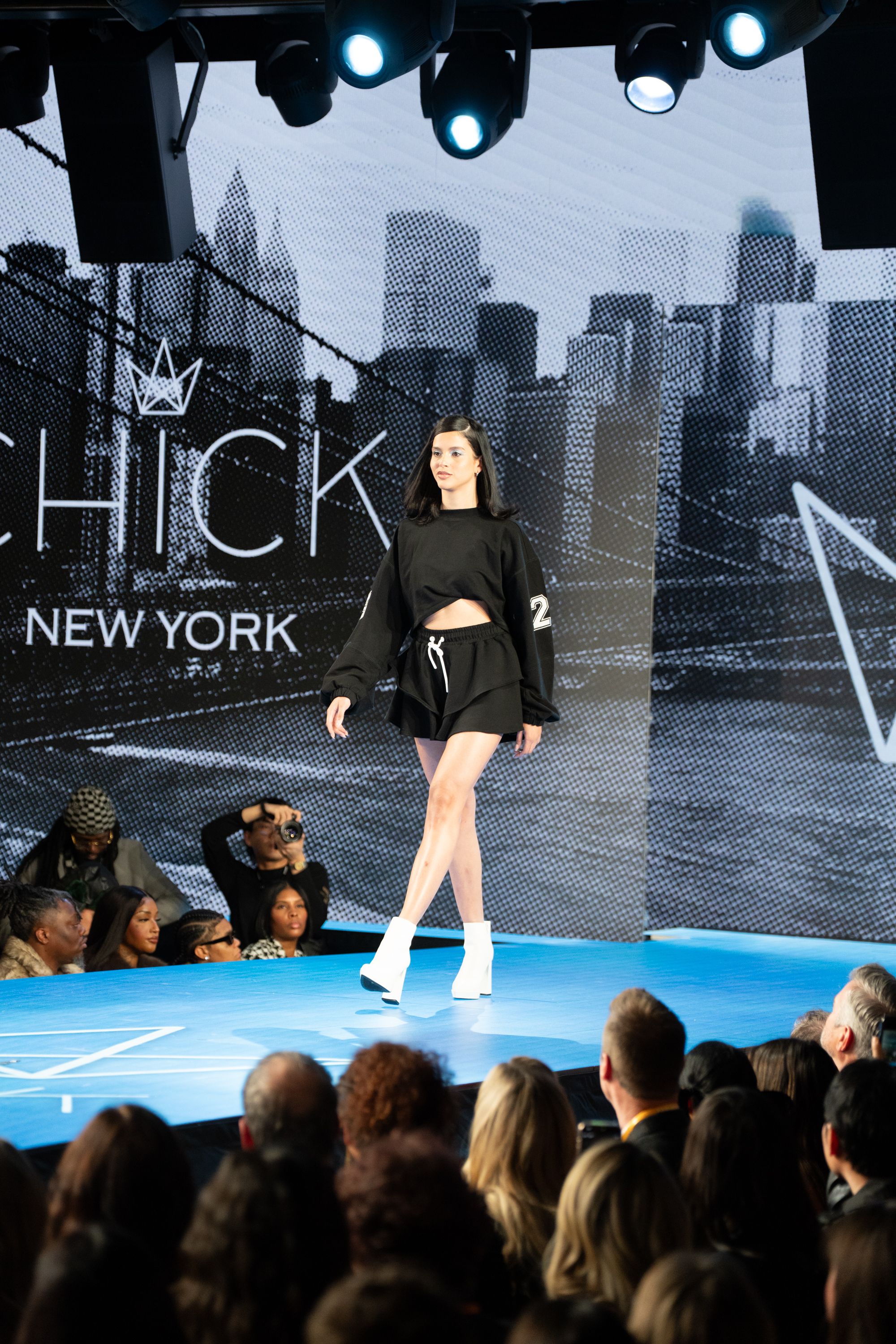 Model walks runway at Chick New York fashion show wearing black crop top and shorts with white boots.
