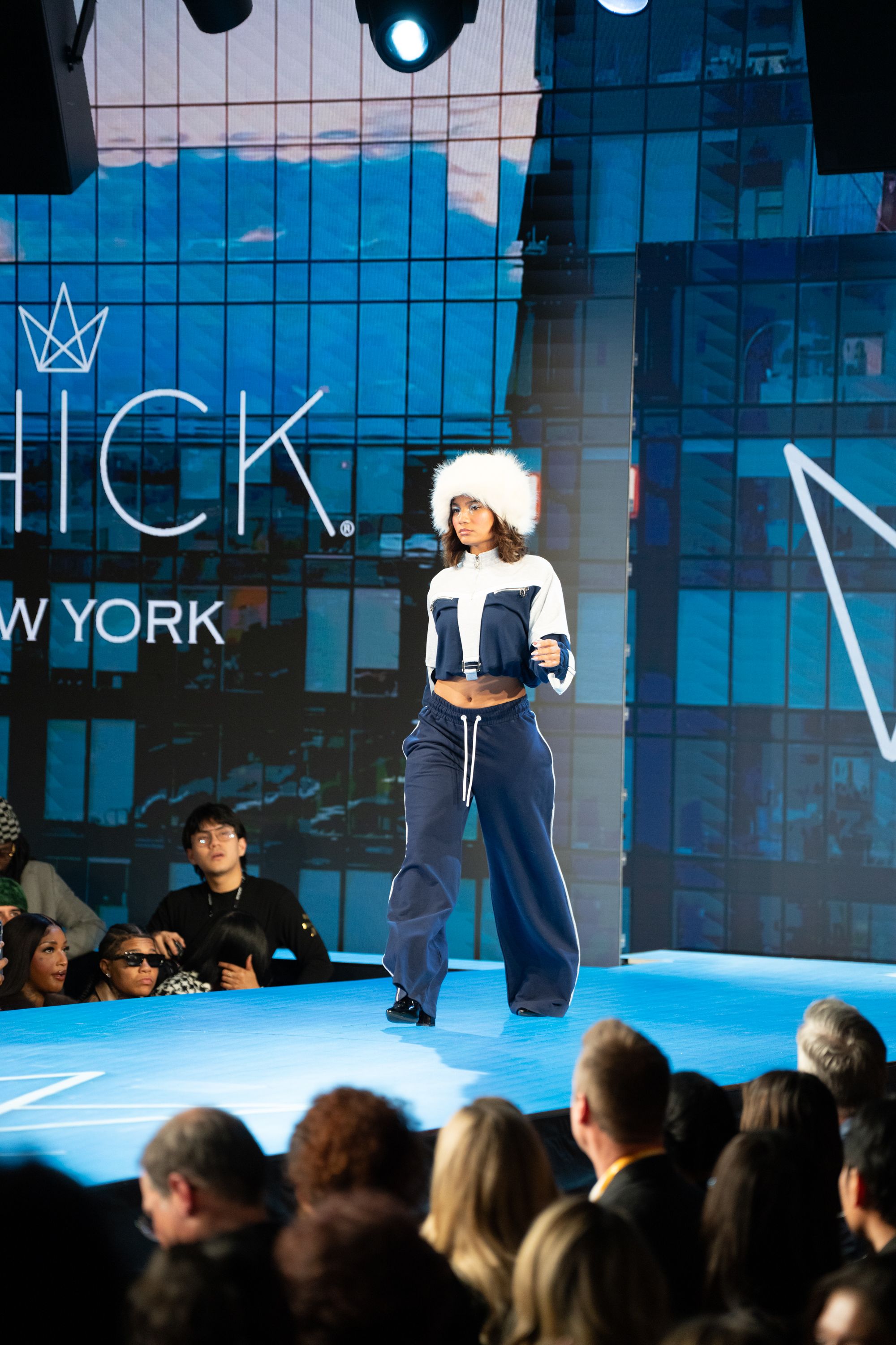 Model walks runway in navy crop jacket, wide-leg pants, and white fur hat at Chick New York fashion show.