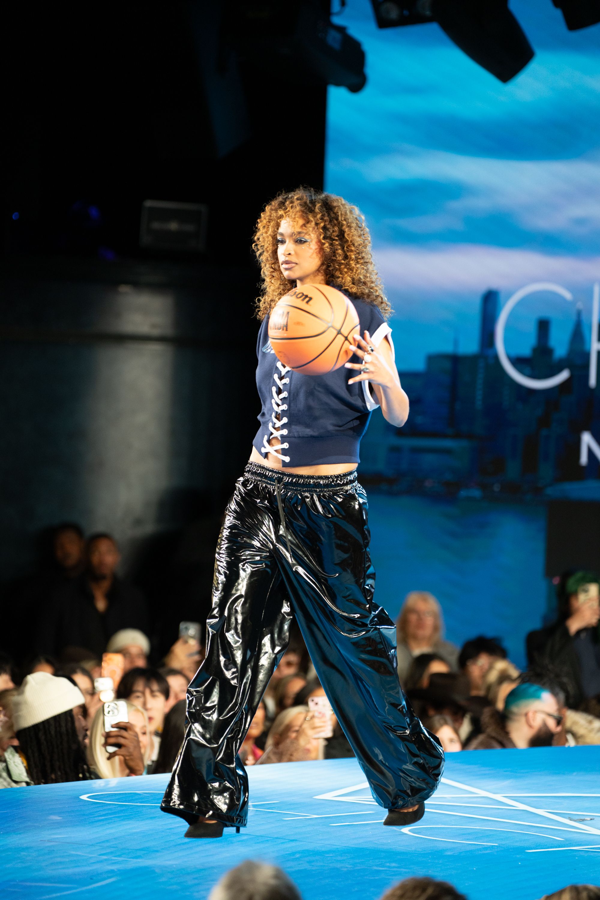 Model walks runway holding basketball in shiny black pants and navy crop top at fashion show.