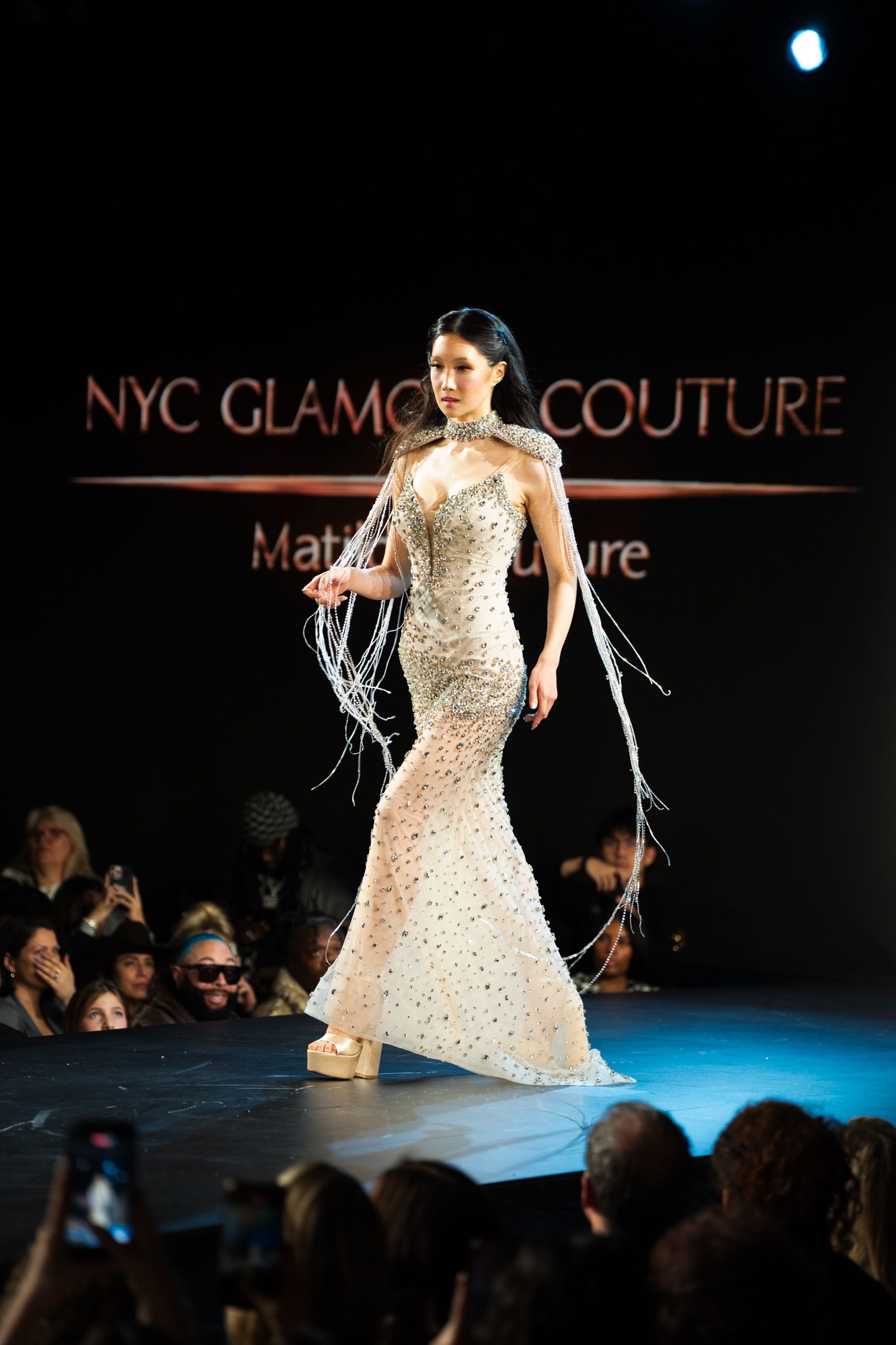 Model walks NYC Glamour Couture runway in beaded mermaid gown with fringe details under dramatic stage lighting.