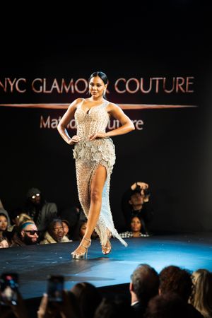 Model walks NYC Glamour Couture runway in elegant beaded gown with thigh-high slit and strappy heels.